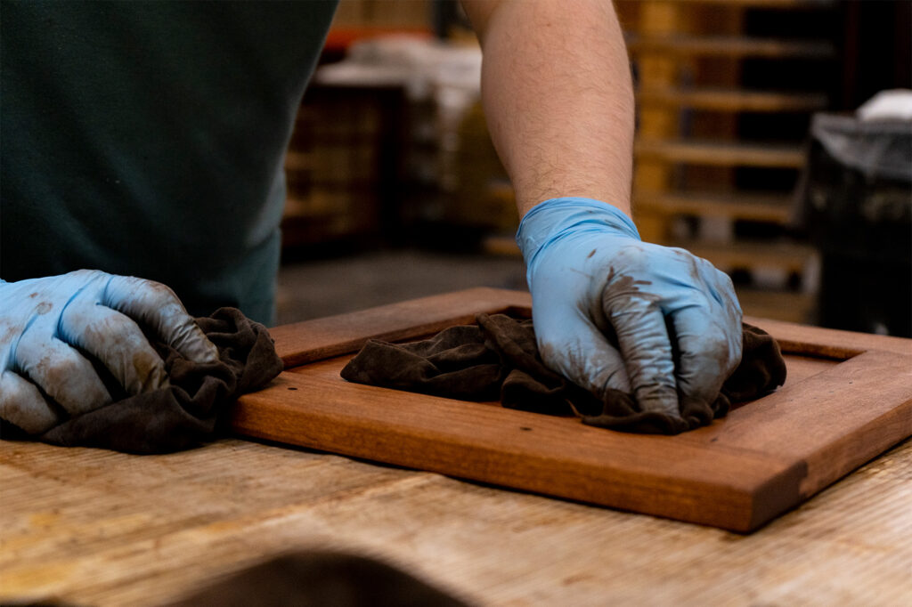 Close-up of a craftsman wearing blue gloves, carefully staining a wooden panel, likely part of a custom jewelry armoire, highlighting the attention to detail in woodworking.